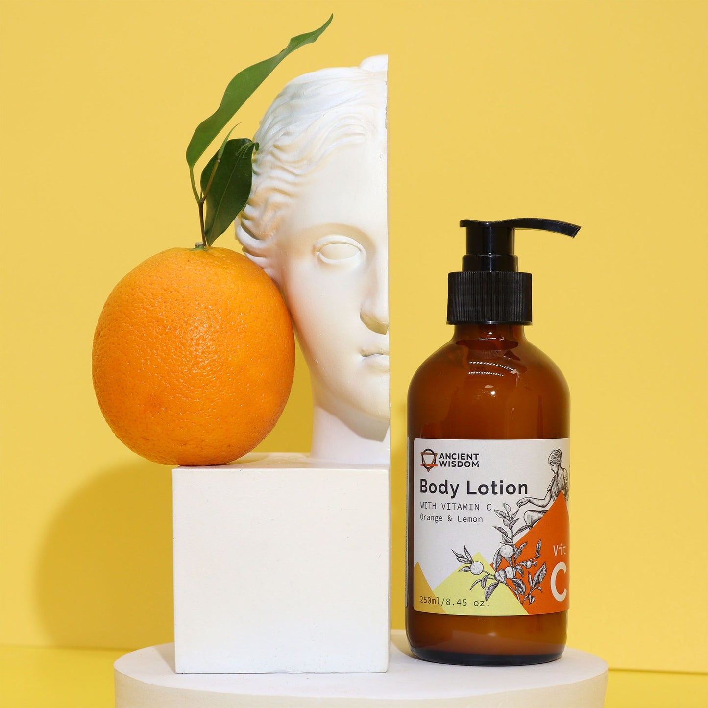 Loción corporal con vitamina C, naranja y limón, en botella con pump, junto a naranja y escultura, fondo amarillo.