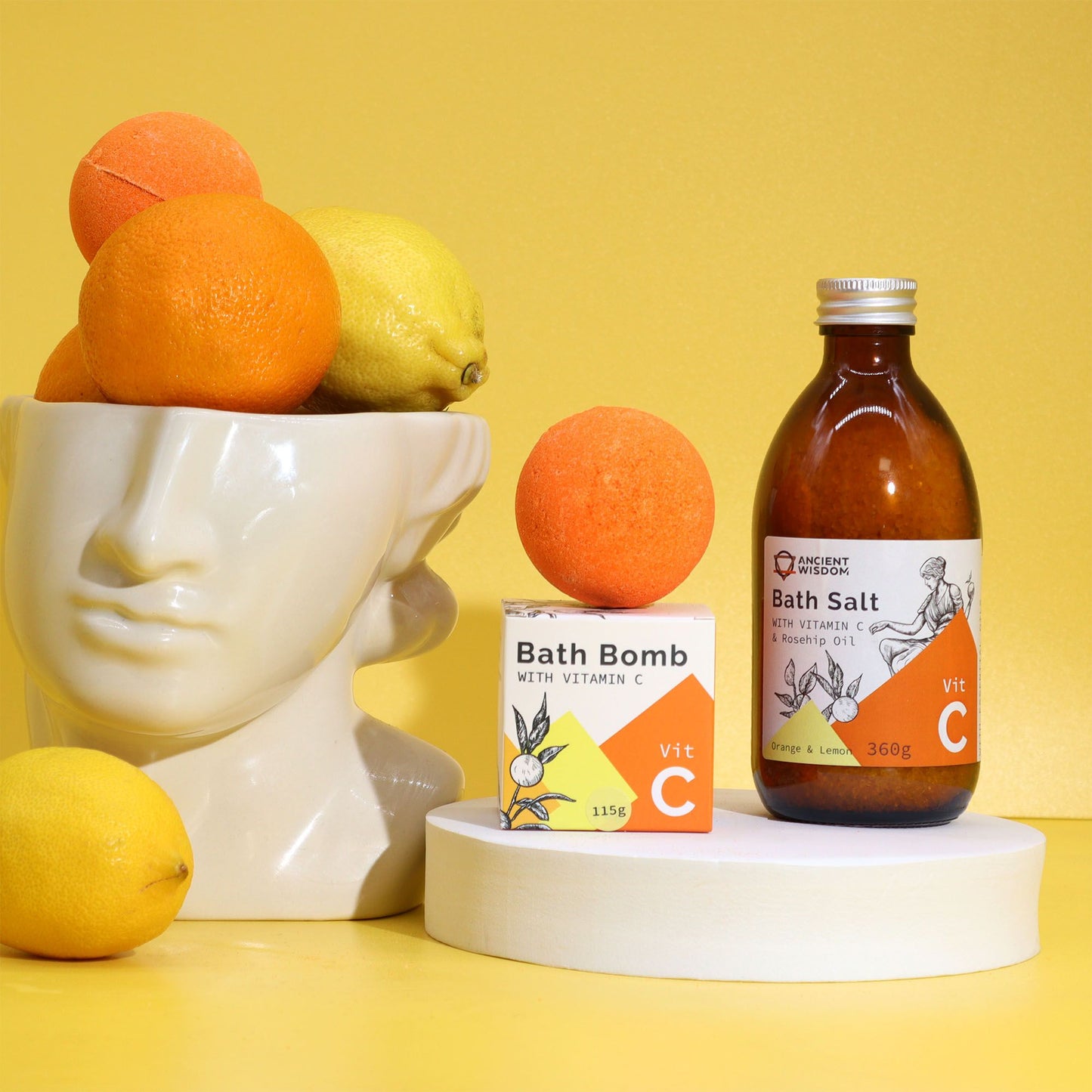 Bomba de Baño con Vitamina C y Aceites Esenciales, caja y frasco con Vitamina C, rodeados de cítricos.