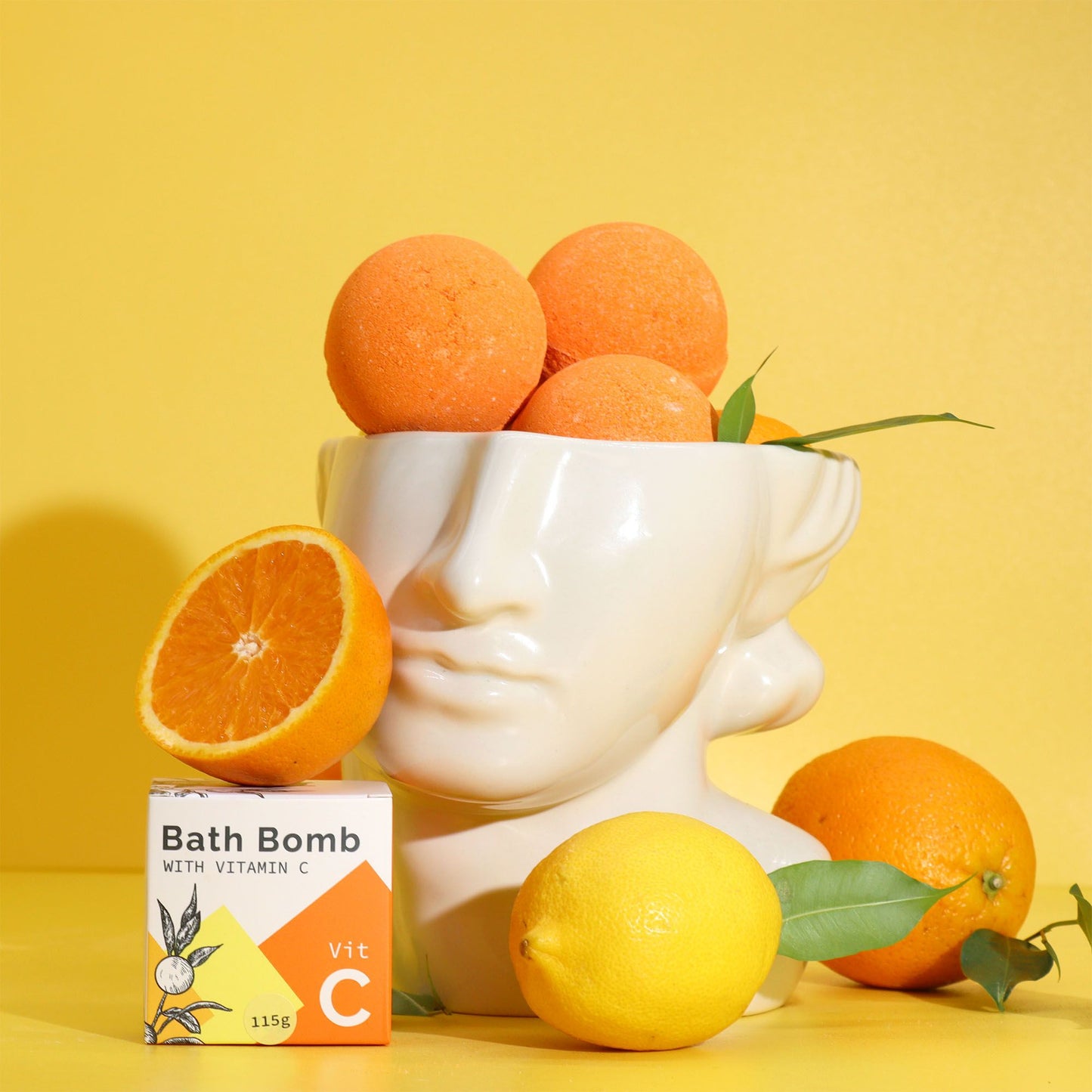 Bombas de baño naranja con Vitamina C y aceites esenciales sobre busto blanco, rodeadas de naranjas.